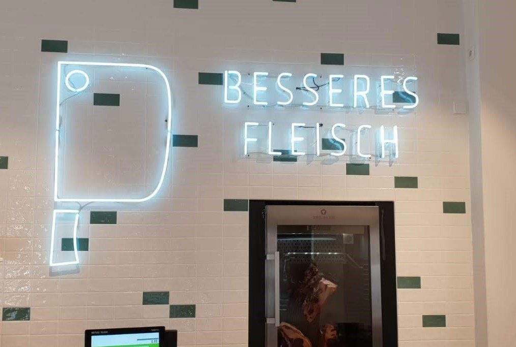 Neonschild für „Besseres Fleisch“ mit der Umrisslinie eines Fleischerbeils über einem Kühlregal im Laden.