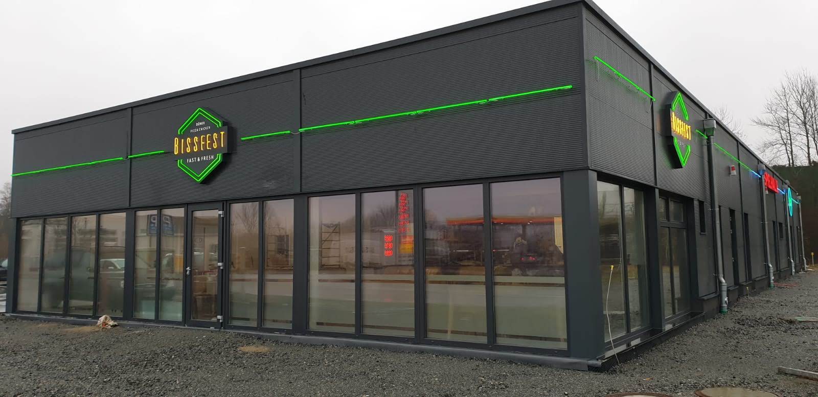 Ein modernes Gebäude mit großen Fenstern, einem grün-schwarzen Logo und einem grünen Streifen oben drauf.