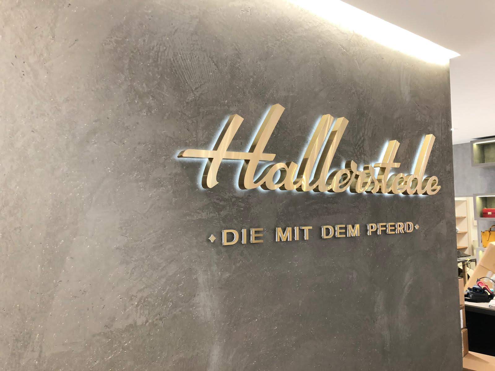 Goldene Schriftzüge auf einer grauen Wand: „Hellerstalle“ und „DIE MIT DEM PFERD“. Angestrahlt durch Deckenbeleuchtung.