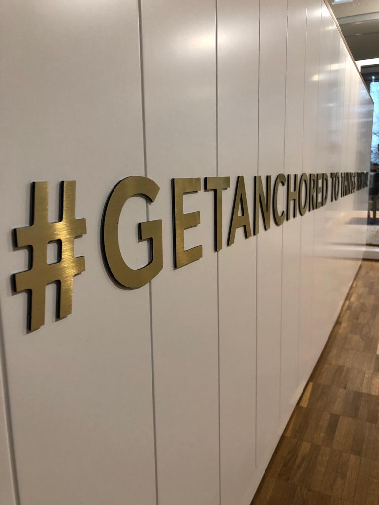 Goldener Hashtag und Text „GET ANCHORED IN HERE“ auf weißer Wand.