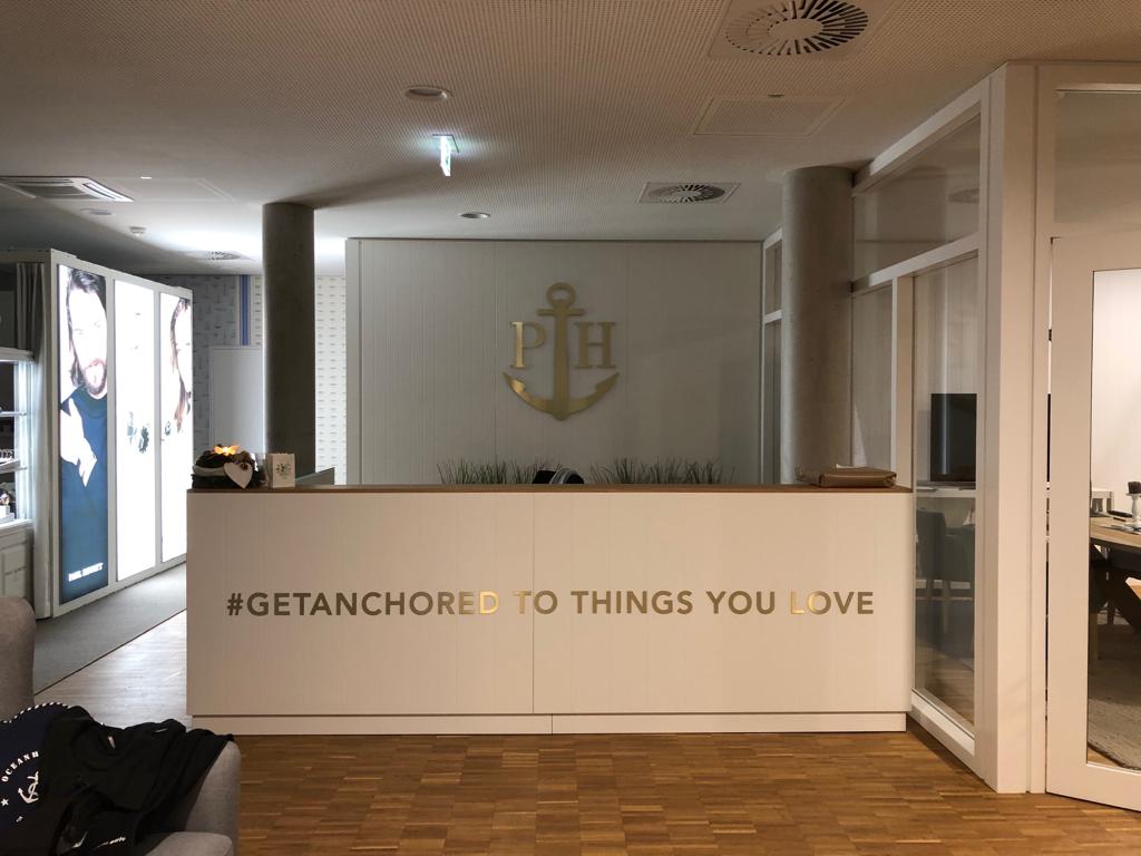 Empfangstresen mit weißer Front, goldenem Anker-Logo, „#GETANCHORED TO THINGS YOU LOVE“ und Holzboden.