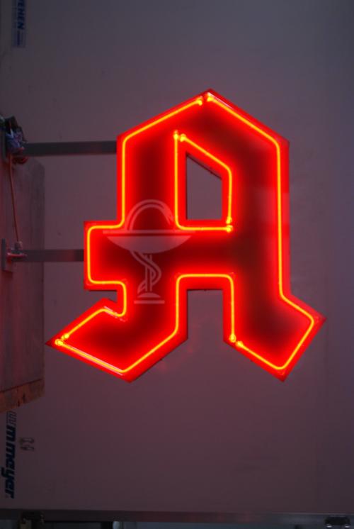 Rotes Neon-„A“ für Apotheke, mit einem Apothekensymbol in der Mitte, auf hellgrauem Hintergrund.