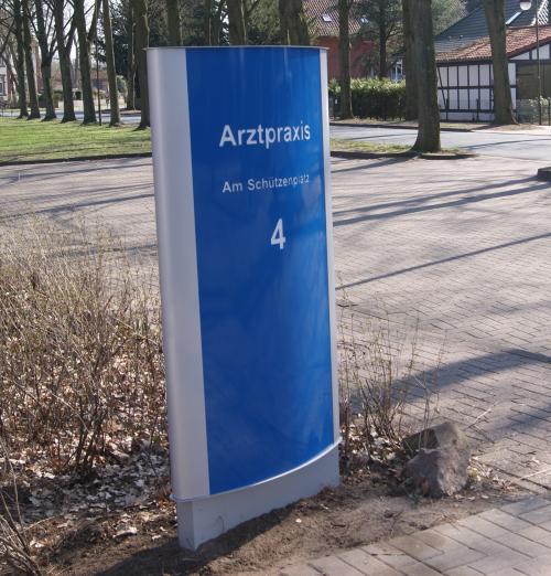 Blau-weißes Schild einer Arztpraxis am „Am Schützenweiher 4“ im Außenbereich.