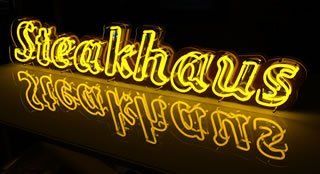 Leuchtreklame: „Steakhaus“ in Gelb, unten auf dunklem Untergrund spiegelnd.
