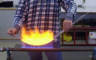 Person im karierten Hemd erhitzt Glasröhre über einer Flamme, Werkstattumgebung.