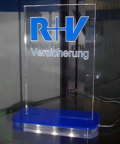 Blau-weißes RHV-Versicherungslogo auf einem klaren Acrylschild mit blauem Sockel.