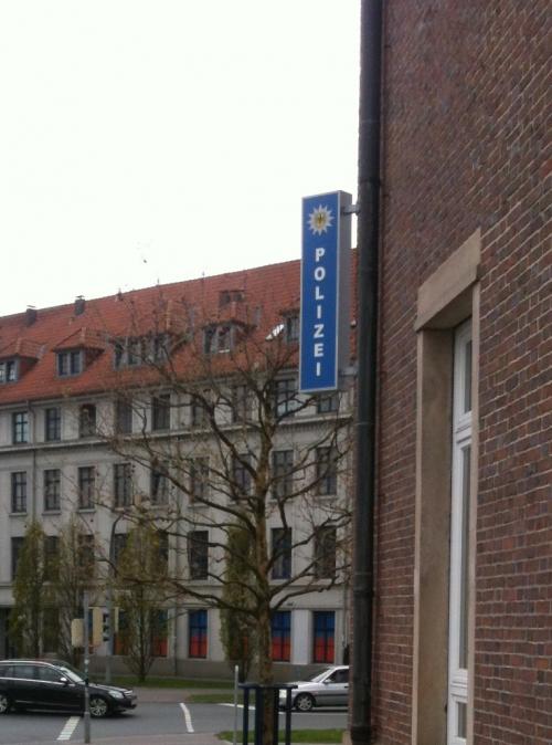 Schild einer Polizeiwache mit der Aufschrift „POLIZEI“ in Blau vor einem Backsteingebäude und einem Gebäude im Hintergrund.