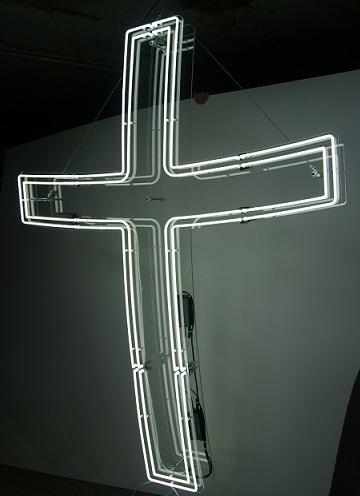 Neonweißes Kreuz hängt an einer grauen Wand.