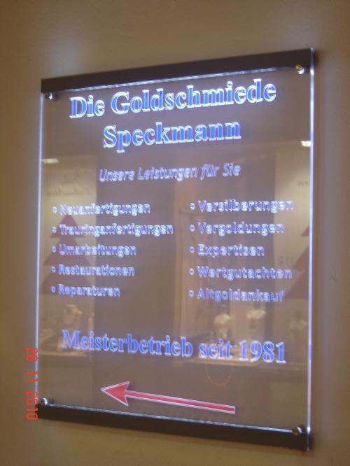 Schild für die Goldschmiede Speckmann. Der Text ist beleuchtet, mit einer Liste der Dienstleistungen und einem roten Pfeil.