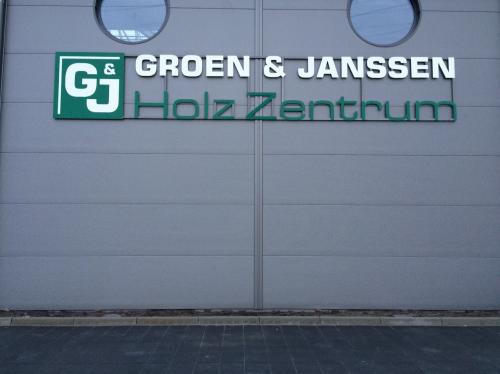 Schild für das Gebäude „Groen & Janssen Holz Zentrum“ mit Logo, grün-weißer Schrift auf grauer Wand.
