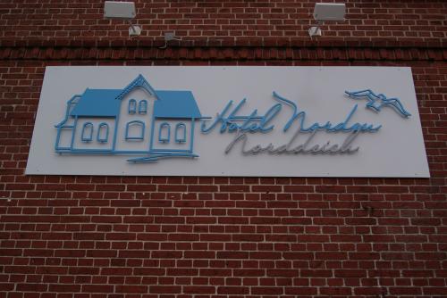 Schild „Hotel Norddeich“ an einer Backsteinmauer; blaues Gebäude und Möwen auf weißem Hintergrund, mit kursiver Schrift.