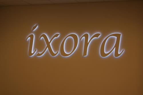 Leuchtend weißes Schild mit der Aufschrift „íxora“ an einer orangefarbenen Wand.