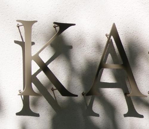 Metallische „KA“-Buchstaben, die an einer weißen Wand angebracht sind und Schatten werfen.