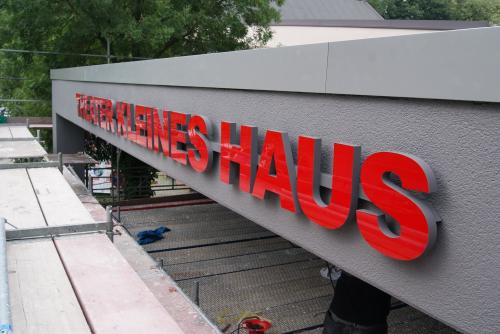Rote Buchstaben „Theater Kleines Haus“ auf einer grauen Gebäudefassade mit Gerüst.