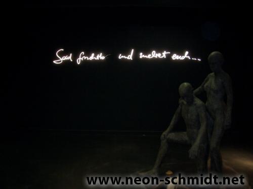 Skulptur aus zwei Figuren unter Neonschrift: „Die Seele findet und trifft dich …“ Dunkle Umgebung.