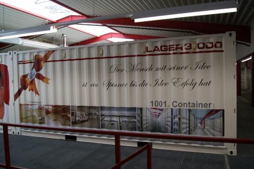 Großer weißer Schiffscontainer mit Werbung, im Innenbereich, vor einem rot-weißen Dach.