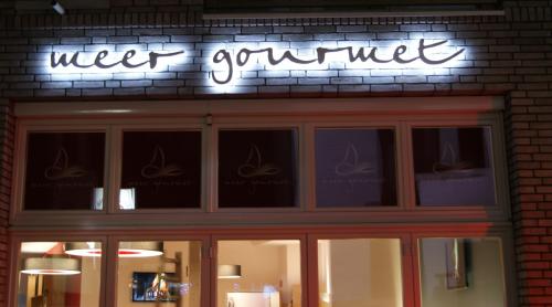 Beleuchtetes „Meer Gourmet“-Schild über einem Restauranteingang mit großen Fenstern.