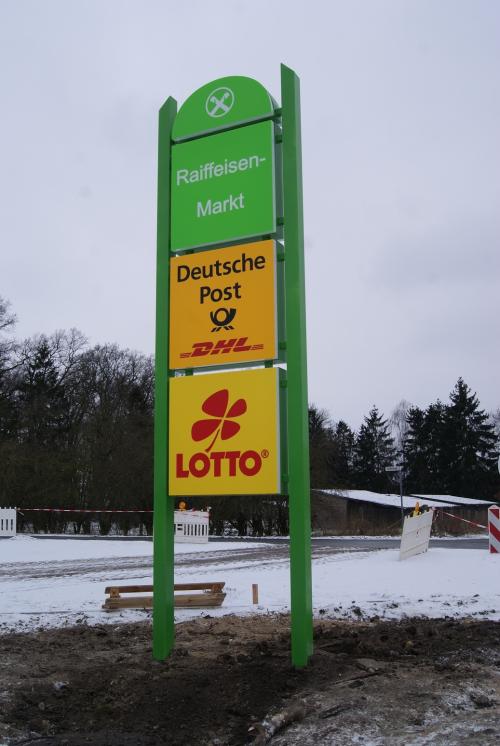 Grüner Wegweiser mit den Logos von Raiffeisen-Markt, Deutsche Post und Lotto vor verschneiter Landschaft.
