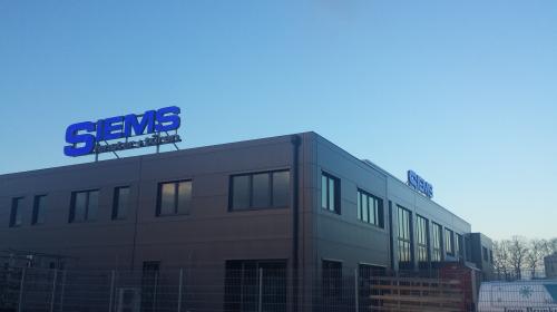 Gebäude mit einem blauen „SHEMS medical+tech“-Schild auf dem Dach, unter einem klaren blauen Himmel.
