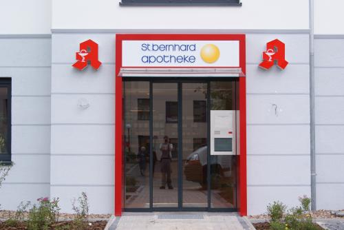 Eingang einer Apotheke mit roten Verzierungen, Glastüren und Apothekensymbolen; Schild „St. Annaro Apotheke“.