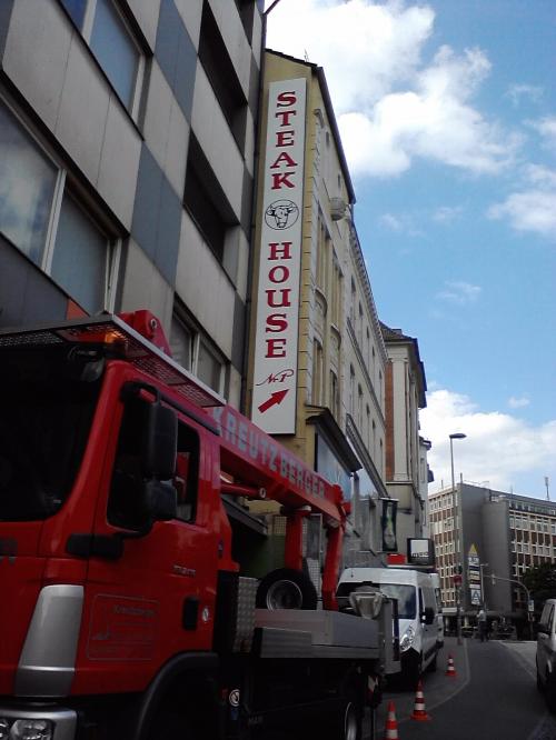 Steakhaus-Schild an der Gebäudefassade, darunter geparkter roter Lastwagen. Schmale Straße, blauer Himmel.