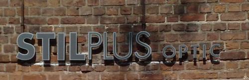 Schild „STILPLUS optic“, metallisch graue Buchstaben auf Ziegelmauerhintergrund.