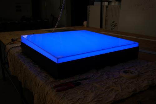 Beleuchtetes blaues quadratisches Display auf einem Tisch. Dunkle Umgebung mit sichtbaren Kabeln und Werkzeugen.