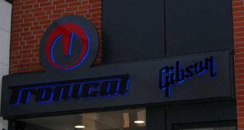 Schild für Tronica Gibson: schwarzer Hintergrund mit blauem LED-Text, rot/blauem Logo und Ziegelwand.