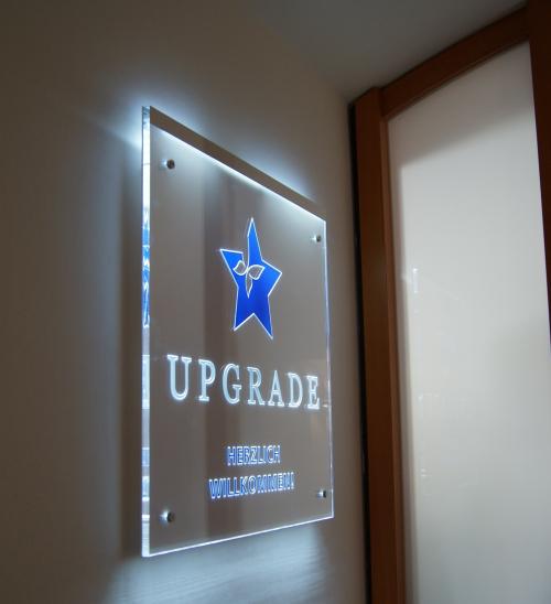 Beleuchtetes Schild für „UPGRADE“ mit einem blauen Sternlogo, an einer Wand in der Nähe einer Tür angebracht.