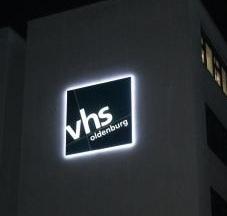 Beleuchtetes „vhs oldenburg“-Logo auf einem Gebäude bei Nacht, quadratisch mit weißem Rand und Buchstaben.
