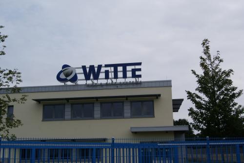 Gebäude mit „WITTE“-Schild, blauem Logo und Zaun. Bewölkter Himmel.