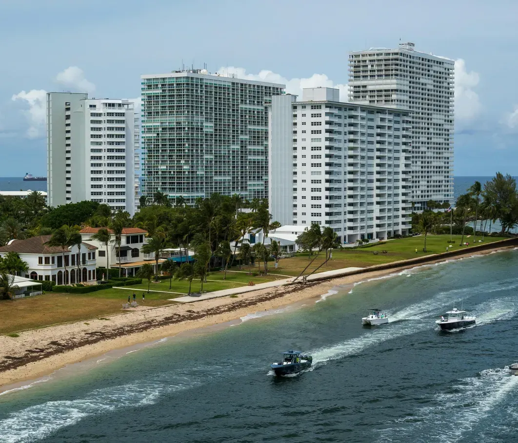 A group of boats are in the water near a beach with tall buildings in the background