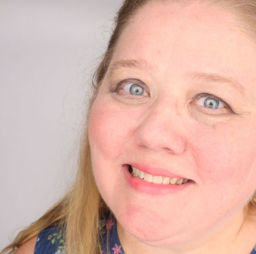 Woman with blue eyes, smiling, looking at the camera; wearing a floral top; light background.