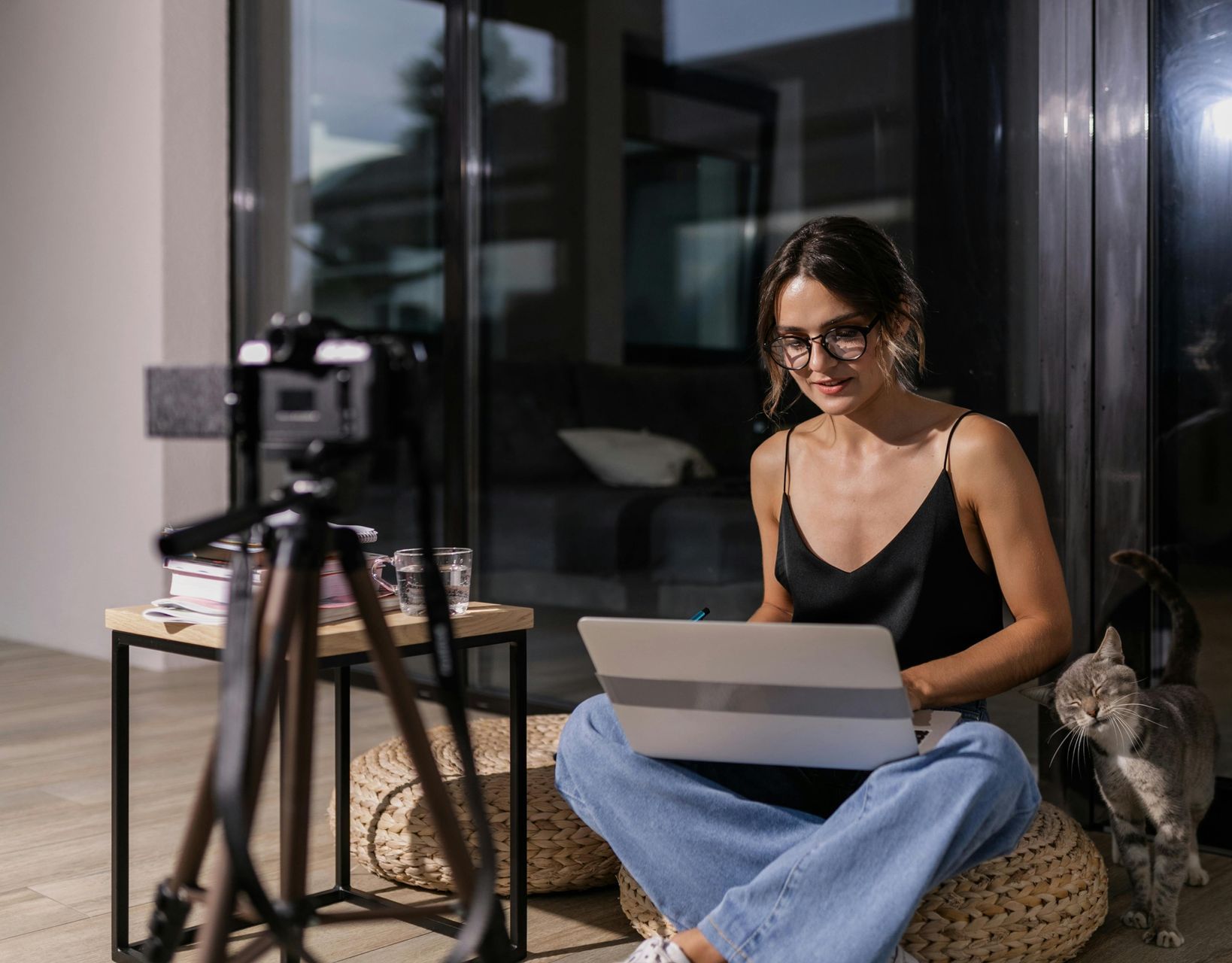 A woman is sitting on a wicker ottoman using a laptop computer.