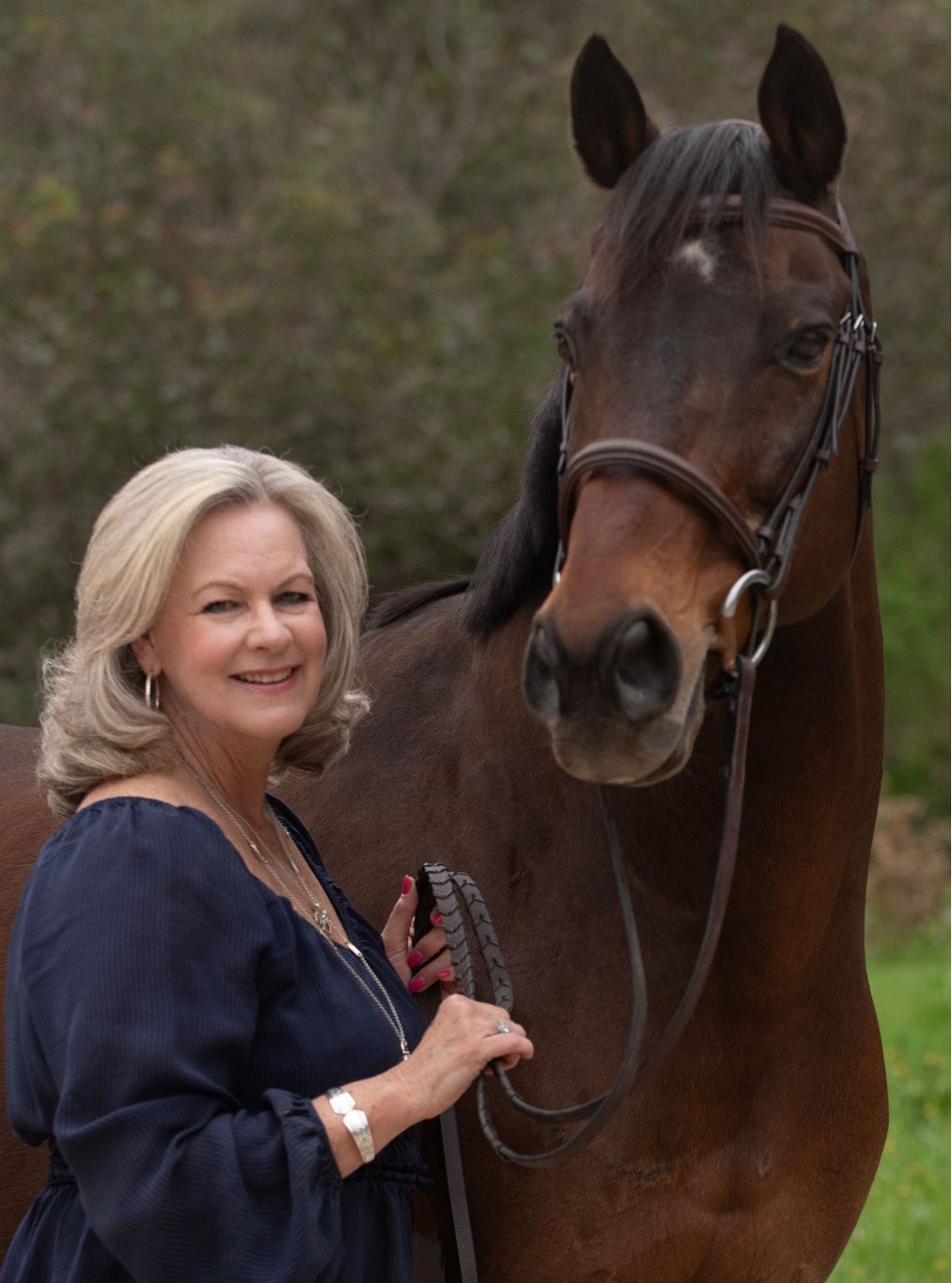 Ann Marie H Woman with blonde hair smiles next to a brown horse wearing a bridle, outdoors.