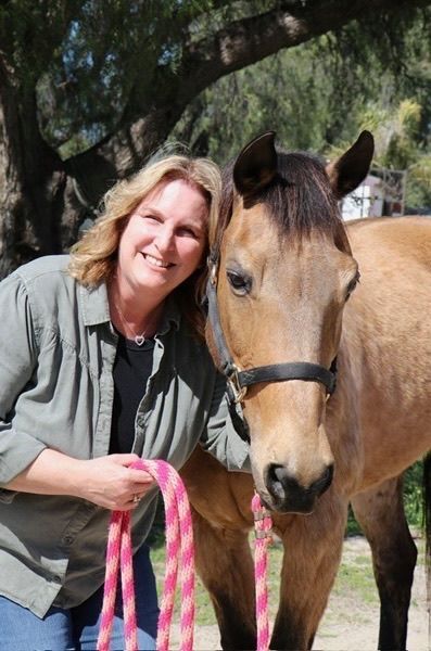 Woman smiles, holding a pink and white lead rope, next to a tan horse wearing a black halter in a sunny outdoor setting.
