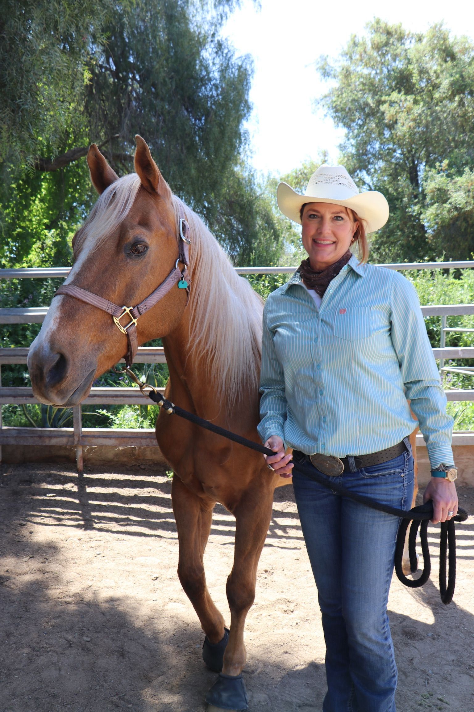 Woman in cowboy hat stands with a palomino horse in an outdoor pen, holding a lead rope.