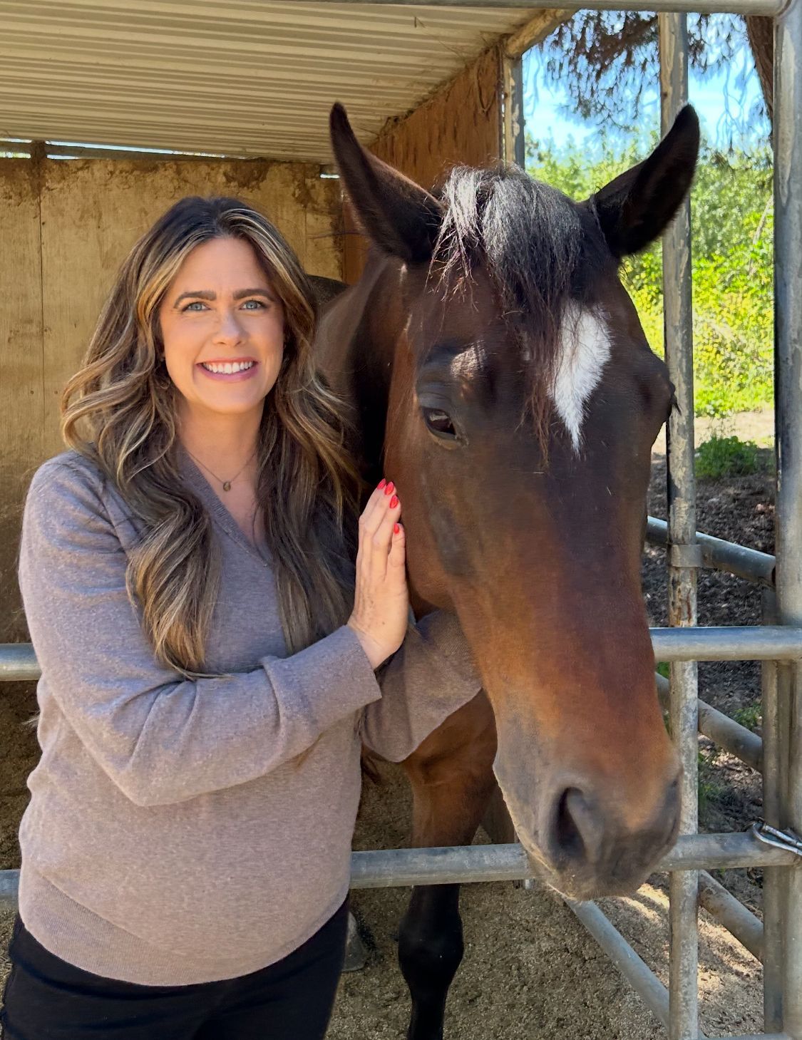 Emily D Woman in a brown sweater smiles, petting a brown horse with a white diamond on its forehead.