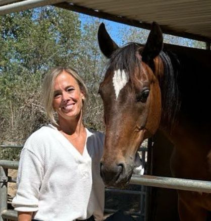 Kelsey N Woman smiles beside a brown horse with a white diamond blaze, outdoors.