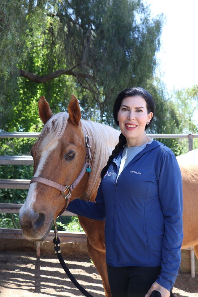 Woman in blue jacket with a tan horse, outdoors in front of a metal fence.