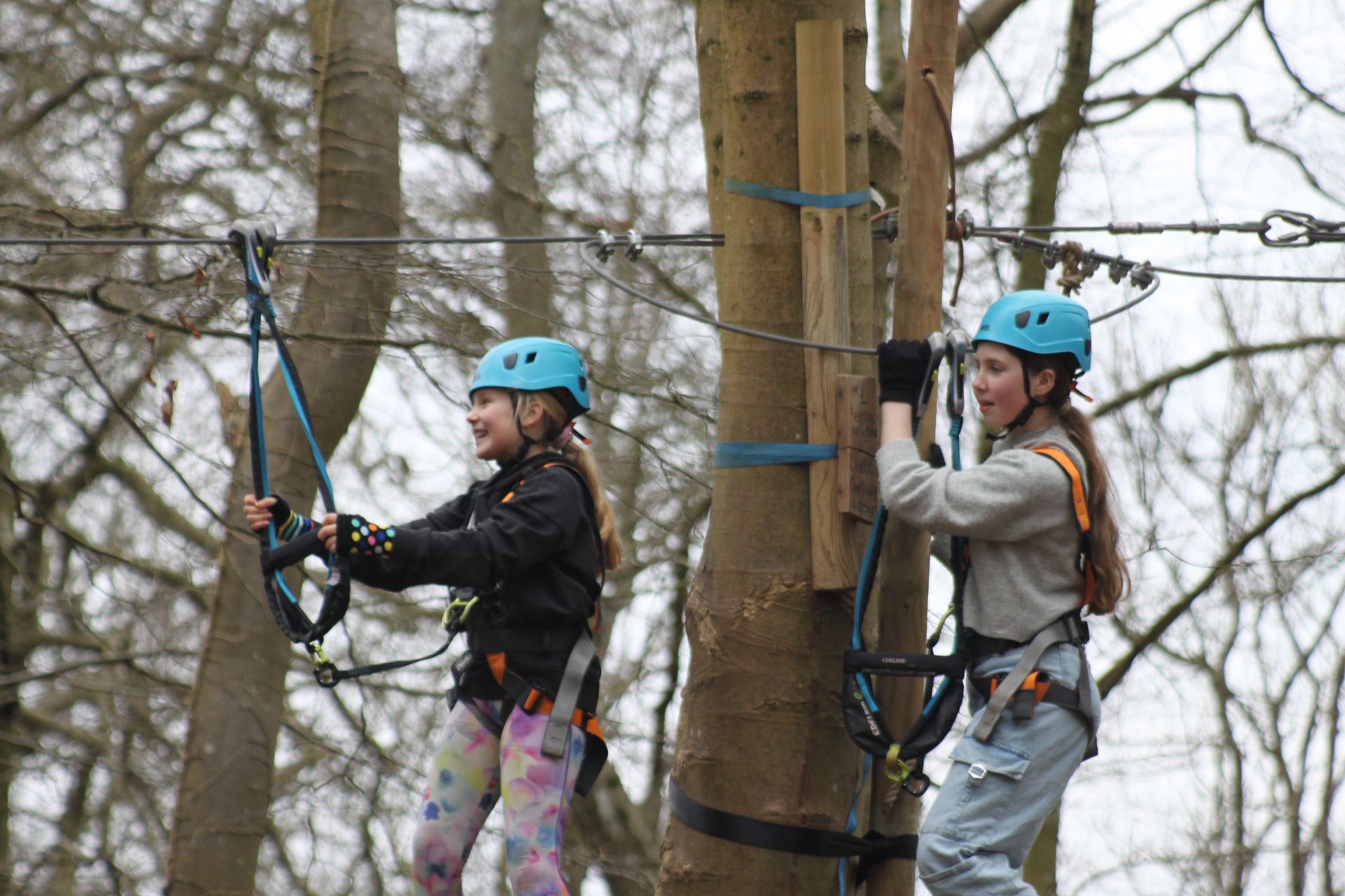 Two girls wearing harnesses and helmets stood in the trees at a birthday party smiling zip wire near me