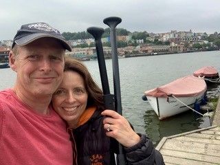Couple near a river holding paddles, smiling. Boats docked nearby, city backdrop.