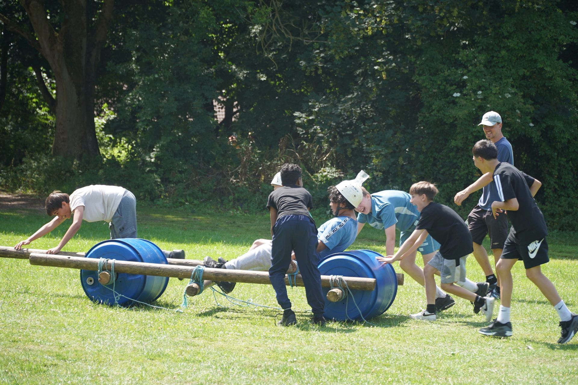 A group of teenage boys smiling pushing a flintstone car made out of wood and barrels whilst on a school trip activity day 
