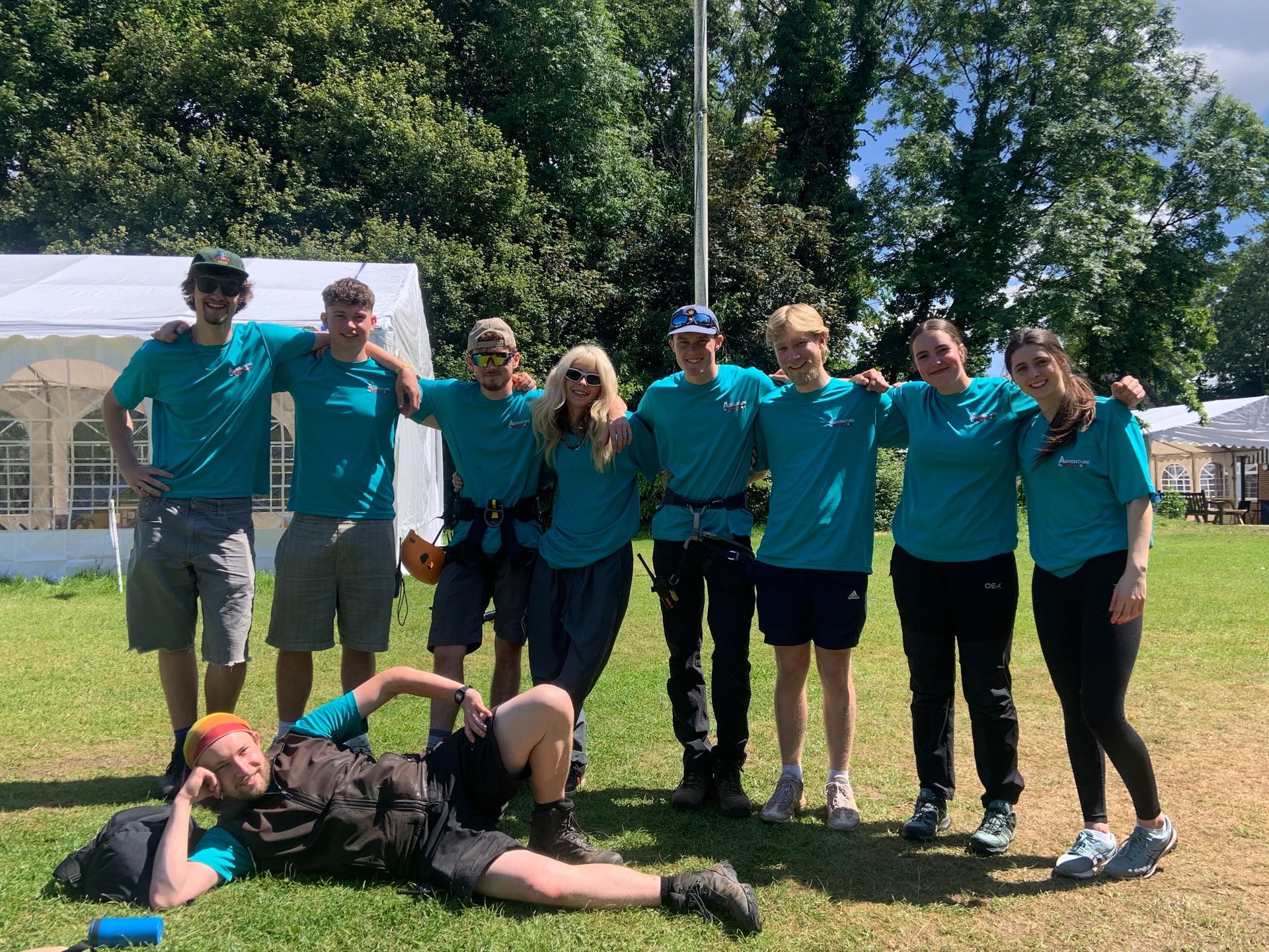 Group of people in teal shirts and shorts posing on grass in front of a white tent. Some have arms around each other.