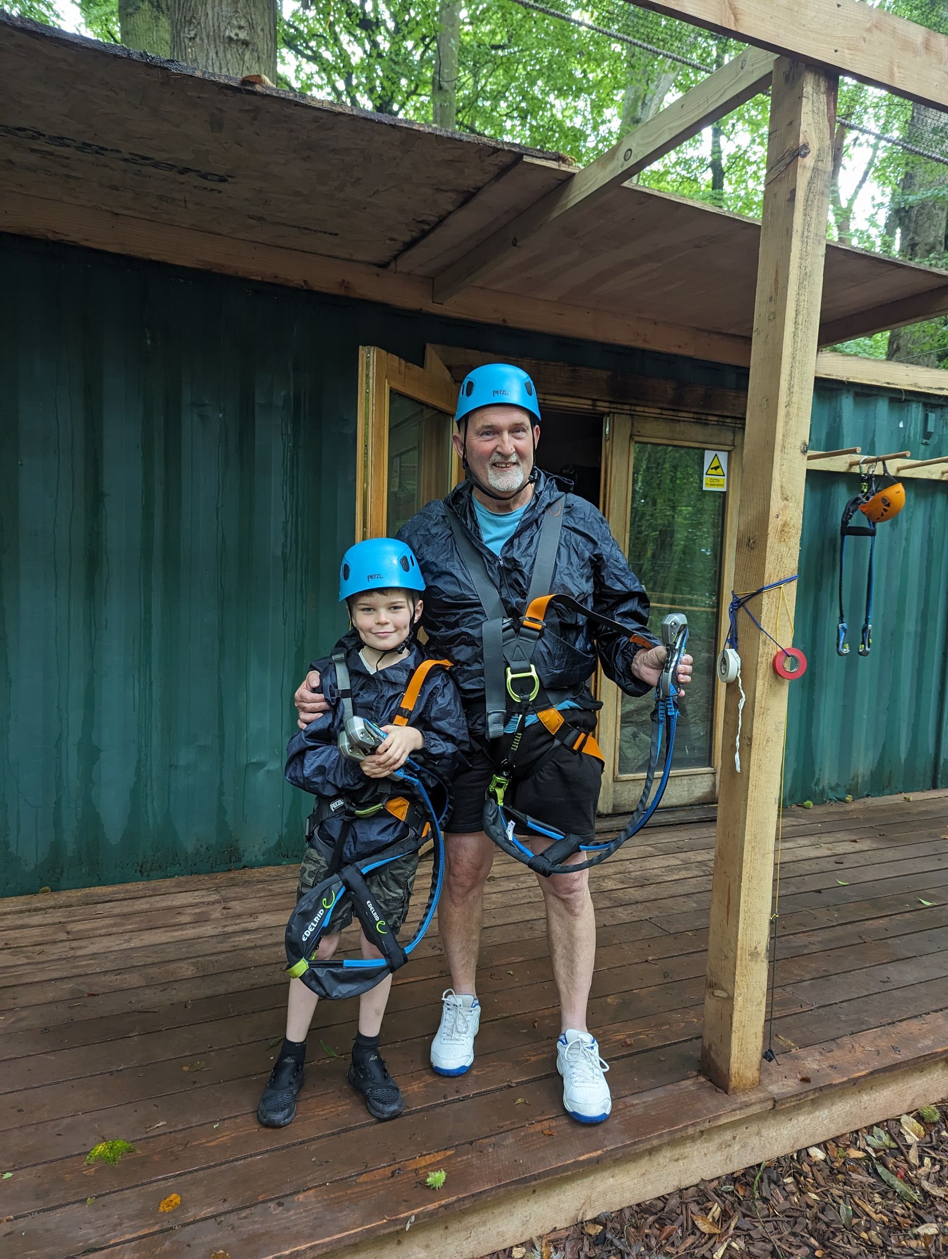 Man and child in climbing harnesses and helmets, standing in front of a green structure, possibly an adventure course.