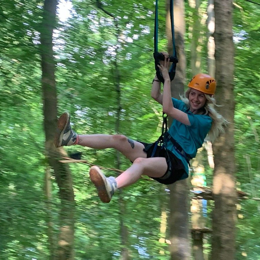 Woman on zip line, smiling, in a forest. Wearing a helmet, harness, and teal shirt. high ropes