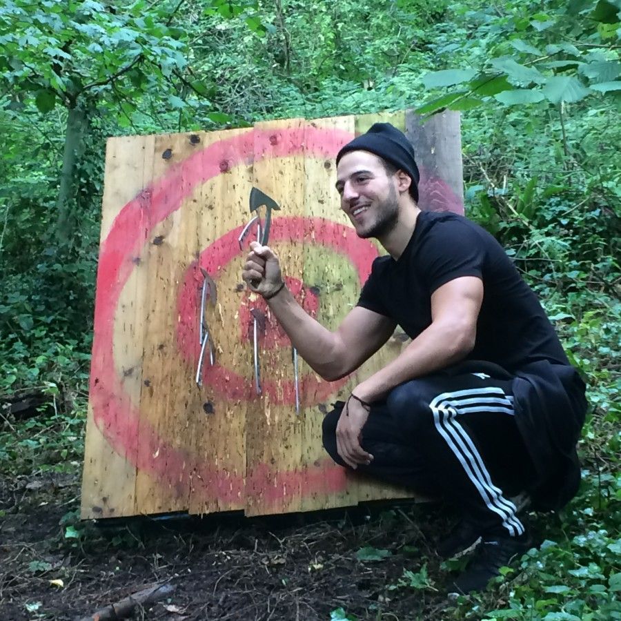 Man squats by axe-throwing target in woods, holding an axe, smiling.