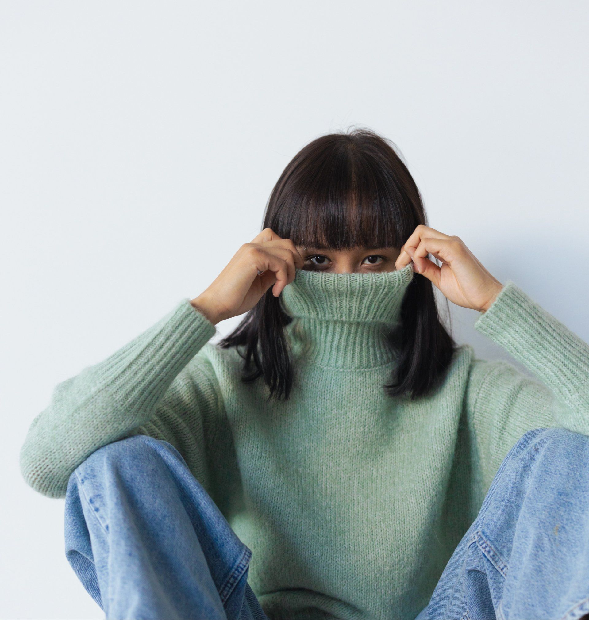 Person pulling up a light green turtleneck. Wearing light blue jeans. Sitting against a white background.