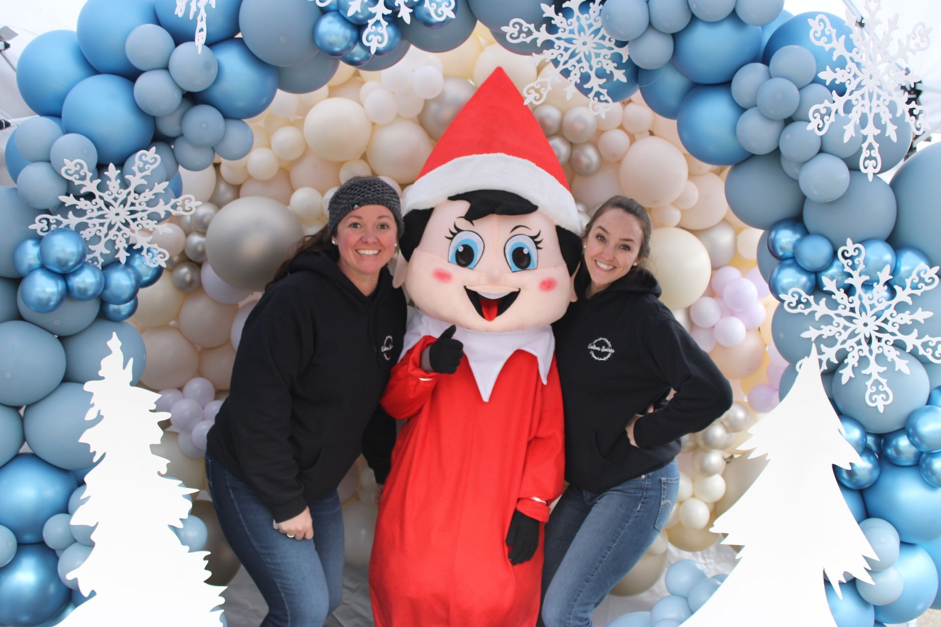 Two women are posing for a picture with an elf on the shelf mascot.