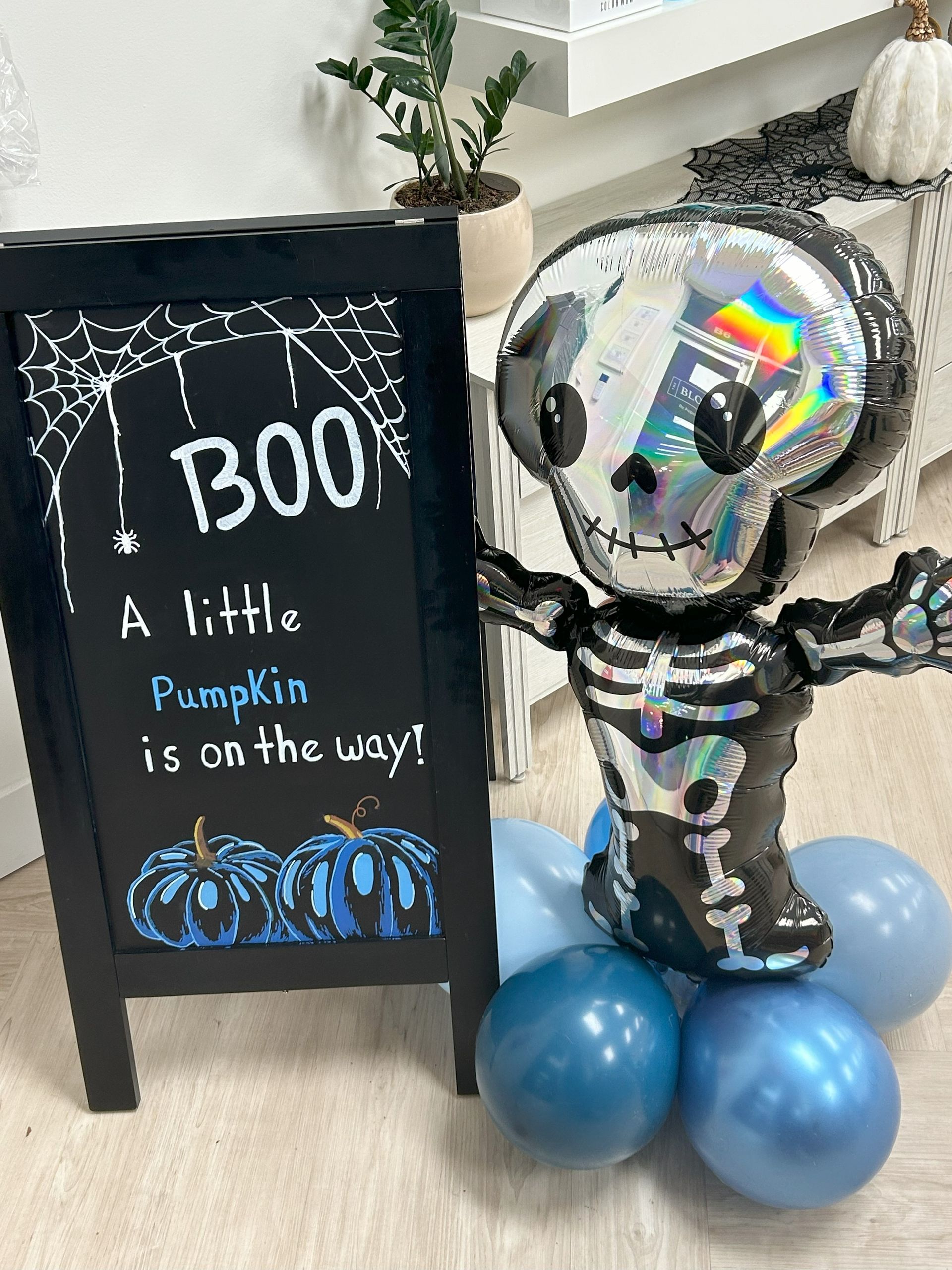 A skeleton balloon is standing next to a sign that says `` a little pumpkin is on the way ''.